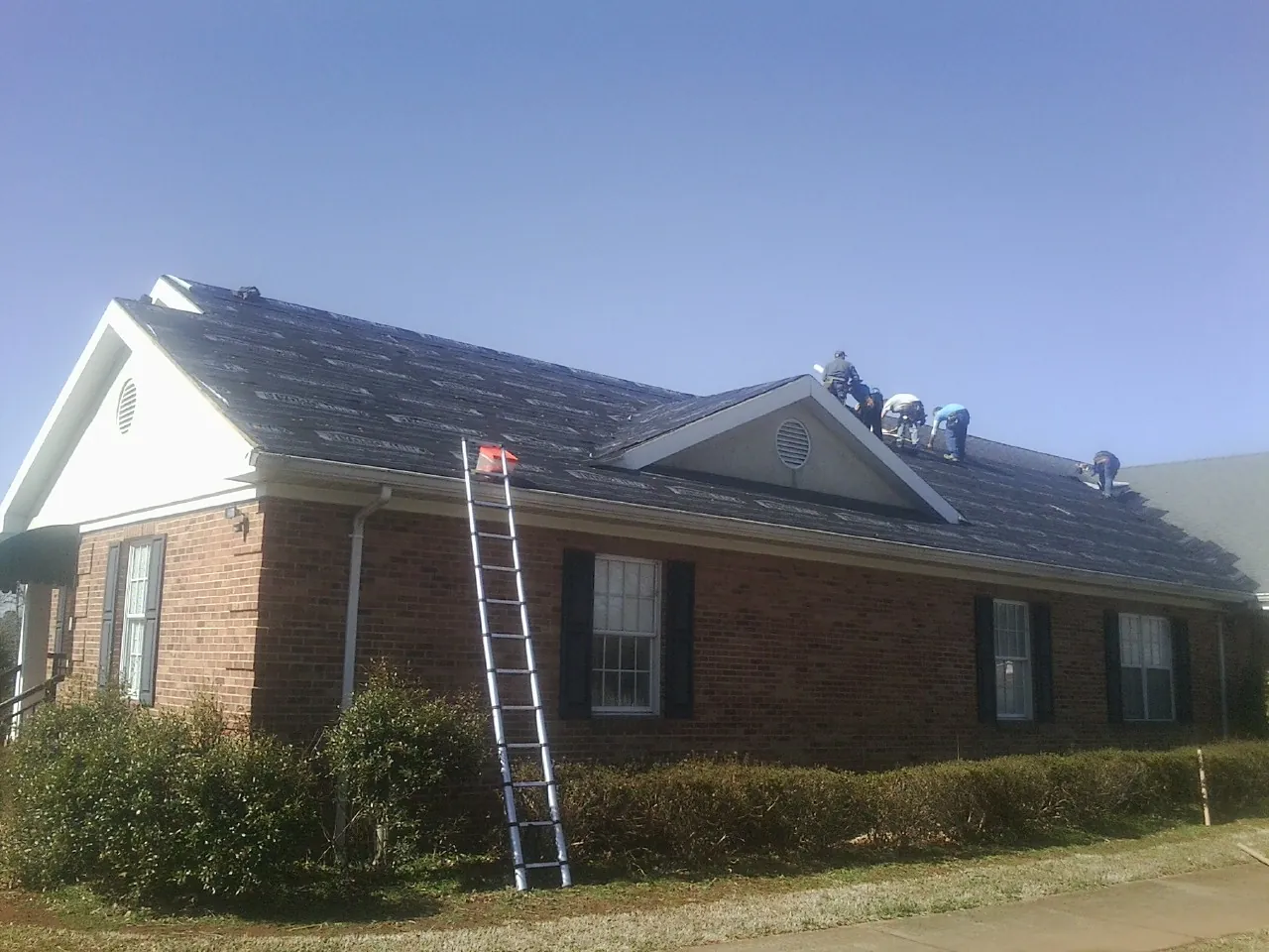 Crew tearing shingles off a brick ranch roof with a ladder set — Emergency Roof Repair work by our crew in Lake Wilson