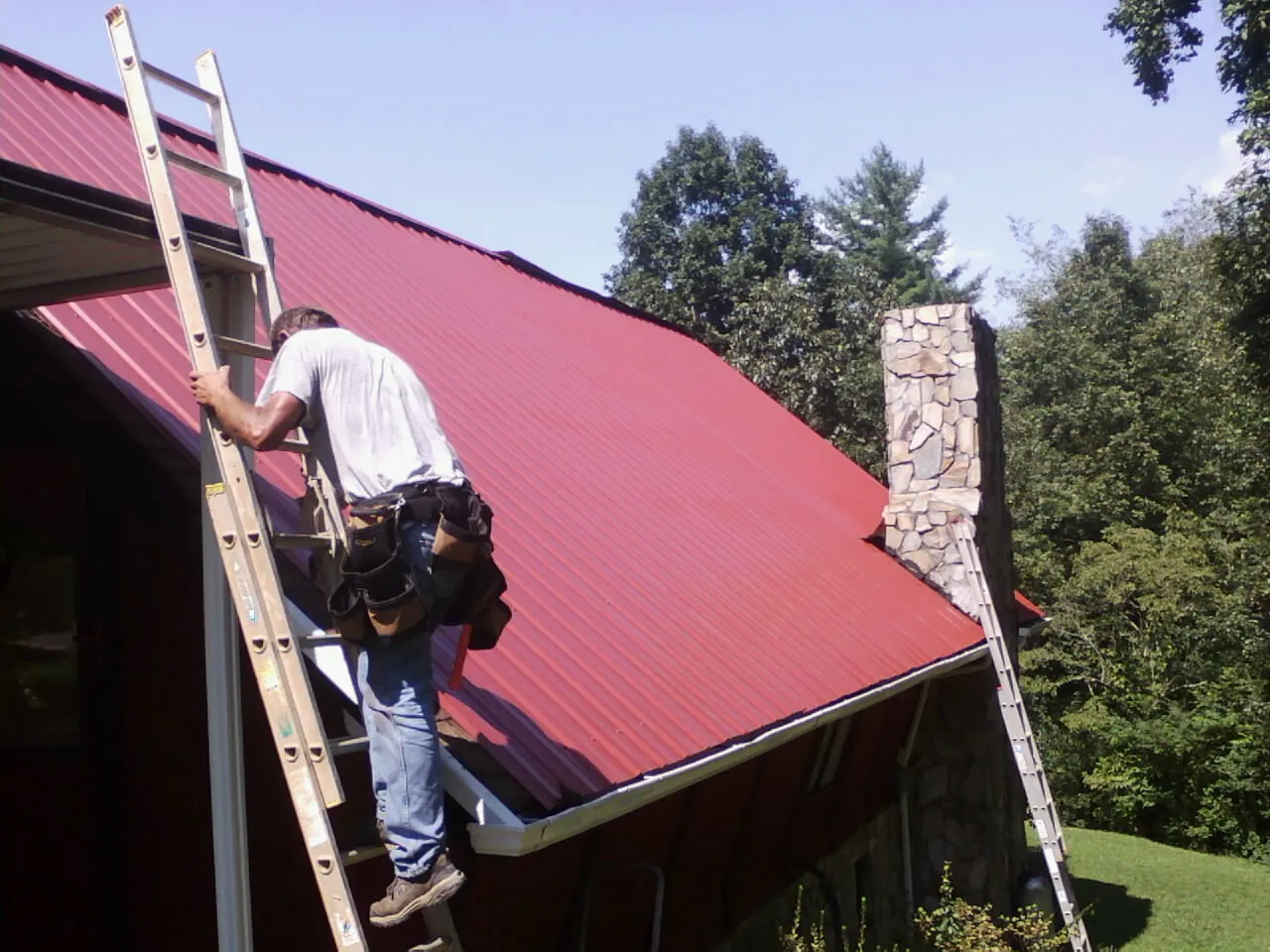 Roofer on a ladder beside a red metal roof and stone chimney — Soffit & Fascia Repair by Roofing Brothers in Lake Wilson